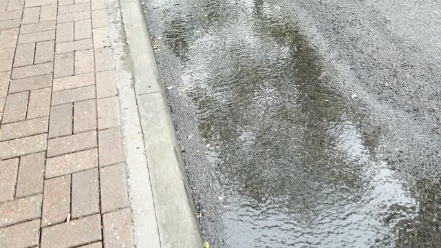 A rippling puddle reflects the sky on a wet asphalt street from a walking point of view. The cool water runs along a concrete curb and neat brick walkway, evoking a calm and fresh feeling.