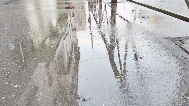 A large puddle pools on a wet asphalt street, functioning like a mirror. The upside-down reflections of buildings and fences create a dreamy, contemplative, and slightly melancholic mood.