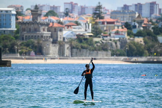 Cascais e Estoril, Portugal - 13 May 2026: View of a man stand-up paddleboarding on calm waters with the historic Palacio do Conde de Castro Guimaraes and city skyline in the background.