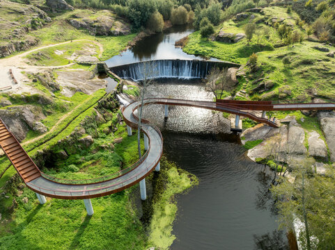 Aerial view of Acude da Ribeira featuring a winding pedestrian bridge and a small dam over the river surrounded by rocky green hills in Ervedal, Coimbra, Portugal.