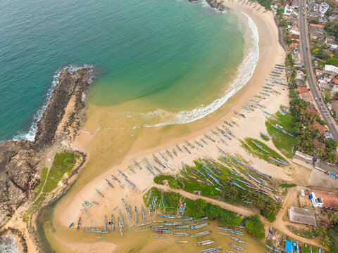 Aerial view of traditional outrigger fishing boats lined up on the sandy beach of a tropical bay with turquoise water and a rocky breakwater Dodanduwa, Southern Province, Sri Lanka.
