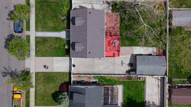 Drone hovers straight down over a lush, landscaped suburban home as a worker tends the grounds below, leaf blower in hand. A bird's eye snapshot of springtime yard care on a sun-drenched day.