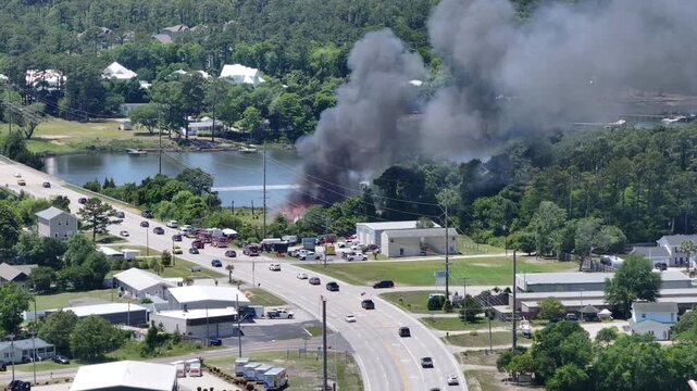 Cinematic aerial drone footage of a residential house fully engulfed in flames during an active structure fire. Thick smoke pours into the sky as emergency lights flicker nearby. Dramatic disaster sce