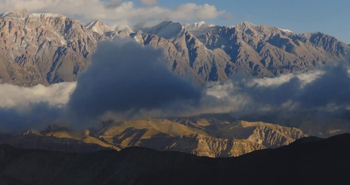 Aerial drone motion moving sideways as thick white clouds drift closely against majestic peaks under a clear blue sky in the remote landscape of Mustang, Nepal.