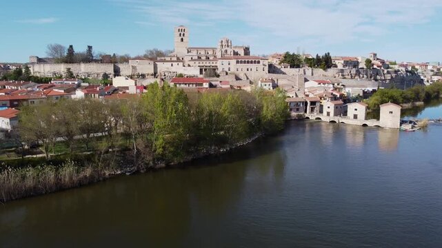 4K aerial drone video ascending from the calm Duero River to reveal the historic skyline of Zamora, Spain. The rising shot captures lush riverside trees, traditional watermills with fishing boats, the