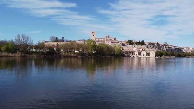 4K aerial drone video advancing over the calm Duero River towards the historic city of Zamora, Spain. The low-angle tracking shot captures the reflection of the medieval skyline, castle, cathedral, an