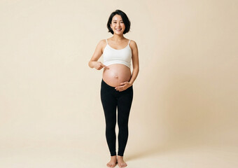 Obraz premium A cheerful pregnant woman with short black hair is smiling at the camera and pointing at her bare belly while wearing a white crop top and black leggings against a plain beige background.