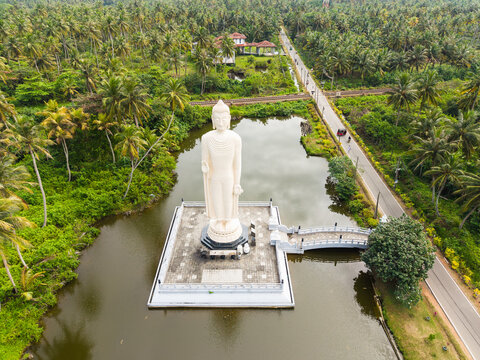 Aerial view of the Tsunami Honganji Viharaya Buddha statue standing in a pond surrounded by lush tropical palm trees and a road in Peraliya, Southern Province, Sri Lanka.