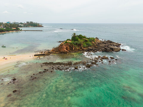 Aerial view of Parrot Rock island surrounded by turquoise ocean water and a sandy beach with people at Mirissa, Southern Province, Sri Lanka.