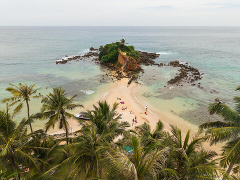 Aerial view of Parrot Rock and the sandy beach with people sunbathing by turquoise waters and palm trees in Mirissa, Southern Province, Sri Lanka.