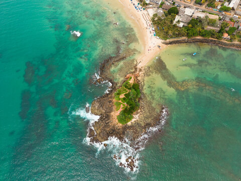 Aerial view of Parrot Rock island connected to the sandy beach by a narrow sandbar surrounded by turquoise ocean water and coral reefs in Mirissa, Southern Province, Sri Lanka.
