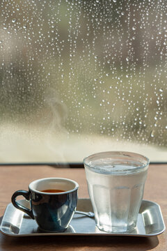 Hot espresso shot with steam and iced water against rainy window.