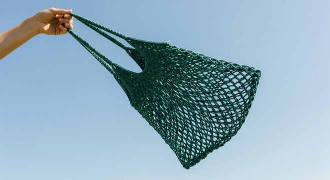 Hand holding a green mesh market bag against a clear blue sky background