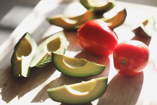 Fresh green avocado sliced and ready to eat along with whole organic roma tomatoes on a cutting board. Delicious healthy, nutritious food.