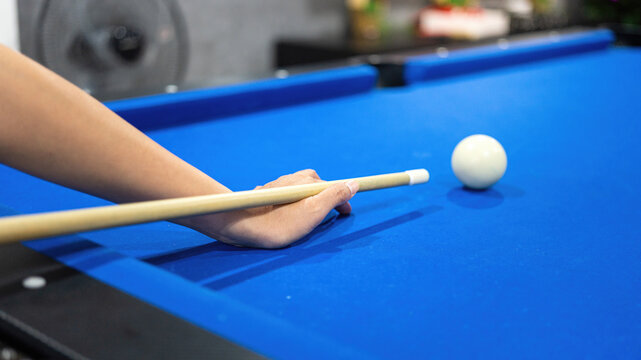 Close-up of a person's hand aiming a cue stick at a white cue ball on a blue billiard table, preparing for a shot with colorful pool balls in the blurred background. Sport action scene.
