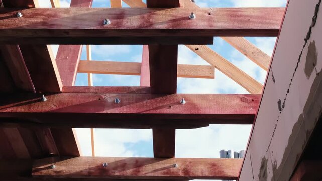 Upward view of wooden roof frame with bolted beams against blue sky, sunlight casting shadows across timber structure, concept of house construction, carpentry work and structural framework
