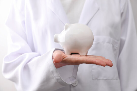 Doctor in medical gown holding piggy bank on white background