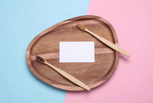 Wooden toothbrushes with white blank business card in wooden tray on pink blue background. Oral care concept. Top view