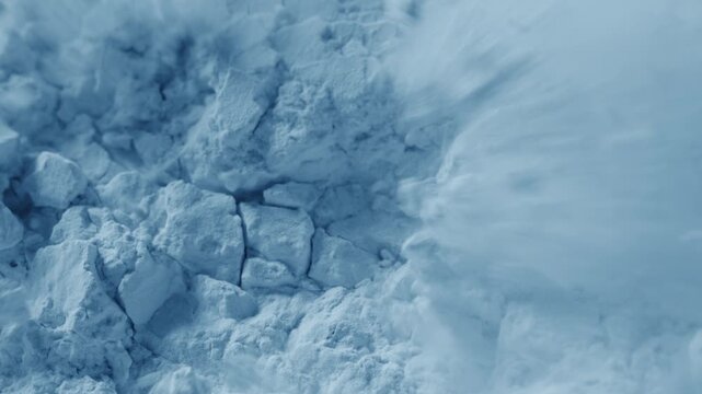 Slow-motion intimate macro view of splintered pale blue powder surface being blown with air stream, breaking its structure and chipping it into small pieces
