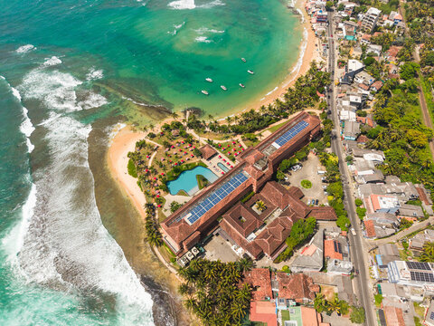 Aerial view of a large beachfront resort with a swimming pool and palm trees along the turquoise coastline in Hikkaduwa, Southern Province, Sri Lanka.