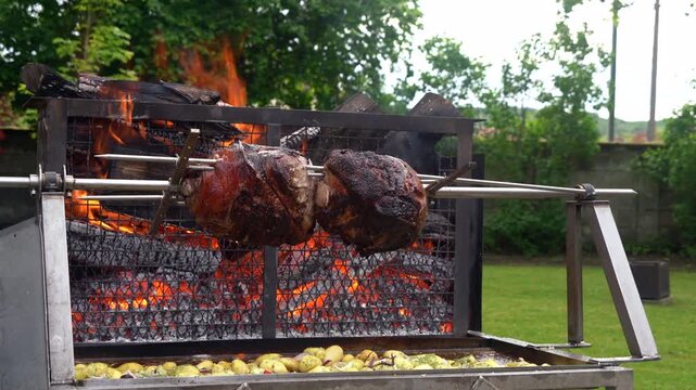 Whole pork ham slowly rotating on a rotisserie spit over open wood fire flames outdoors in France. Potatoes cook below collecting meat drippings at a traditional Frenchfeast similar to mechoui