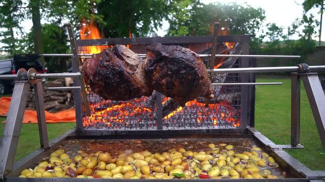 Whole pork ham slowly rotating on a rotisserie spit over open wood fire flames outdoors in France. Potatoes cook below collecting meat drippings at a traditional Frenchfeast similar to mechoui