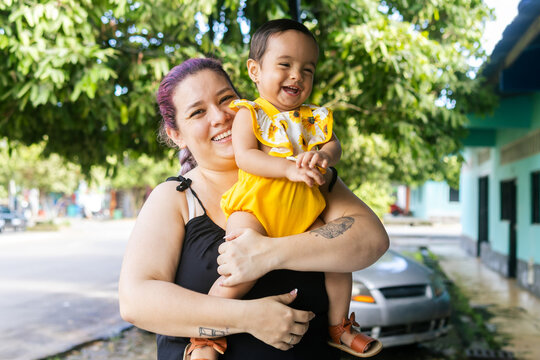 A Latina mother holding her smiling baby outdoors on a residential street, reflecting love, happiness and family bonding, adult Latina woman and baby 1 to 2 years old.