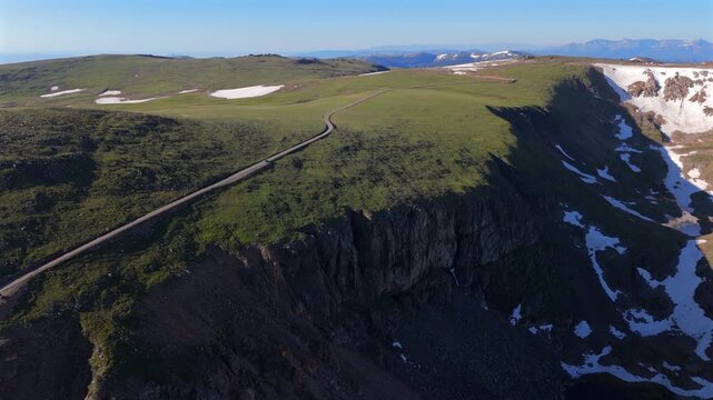 Aerial of Beartooth Highway in Montana, USA showing winding mountain road along dramatic cliff edge with green alpine tundra, snow patches, and distant peaks under clear blue sky.