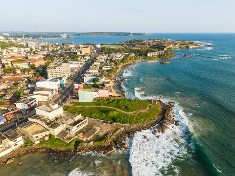 Aerial view of the coastal city with urban buildings and turquoise ocean waves crashing on the shore near the historic Galle Fort in Galle, Southern Province, Sri Lanka.
