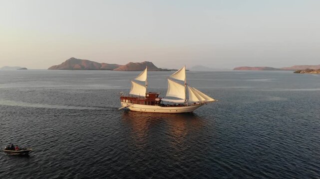 Aerial drone view of a traditional Indonesian wooden Phinisi boat sailing with full white sails during sunset in Labuan Bajo, Komodo National Park
