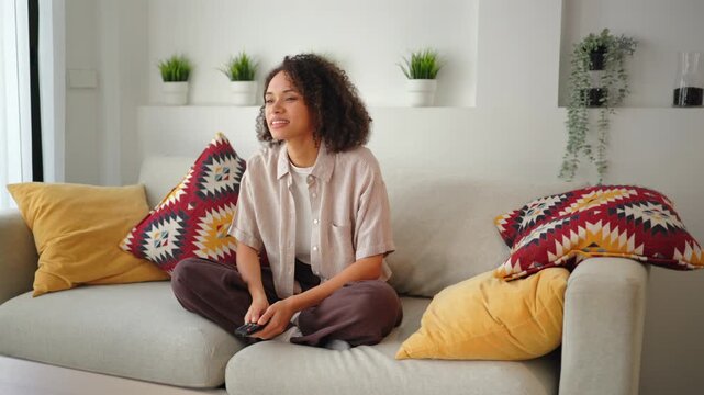Young African American woman with curly hair on sofa, smiling while changing TV channels with remote. Relaxed entertainment lifestyle in a bright modern living room with colorful pillows.