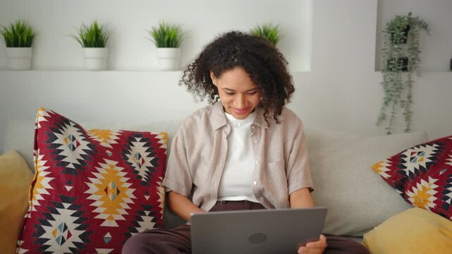 Young African American woman with curly hair sitting cross-legged on sofa, using a laptop. Remote work lifestyle in a bright modern living room with colorful ethnic pillows and plants.