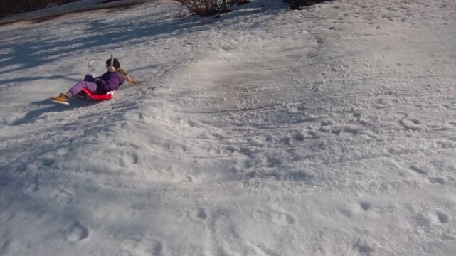Handheld tracking shot follows a young child sliding down a snowy hillside using a plastic shopping bag as a makeshift sled. Sunlight gleams across the snow terrain at Budapest Normafa park.