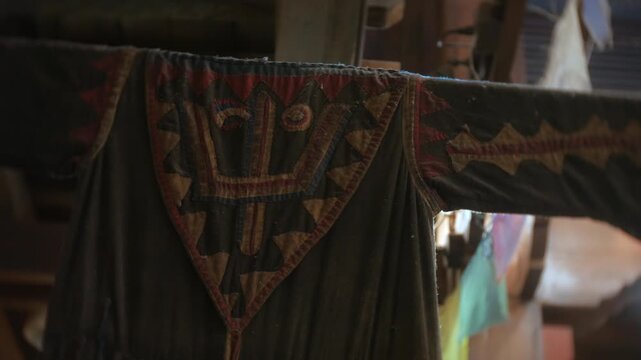 Textile clothing hangs inside a Tibetan shrine, showing worn black fabric with red and tan stitched patterns. Close-up view highlights handmade ritual decoration, soft indoor light, and aged texture