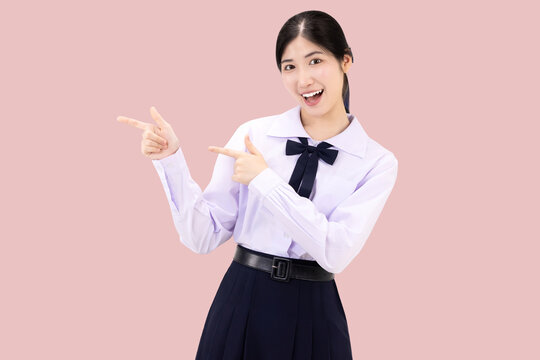 Excited Asian high school student girl in Thai school uniform pointing fingers at copy space for advertising, smiling happily, isolated on pink background.