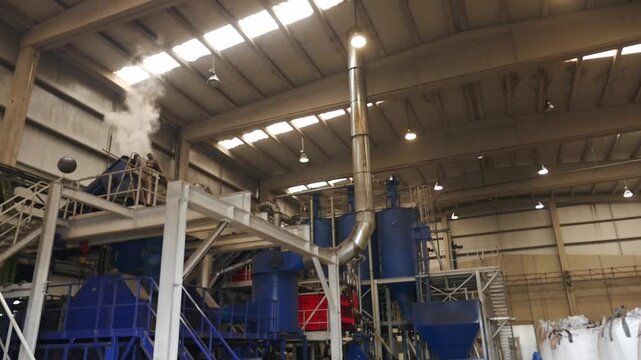 Used rubber recycling factory, revealing heavy blue machinery, industrial ladders and a metal pipe emitting white steam under the roof of a warehouse.