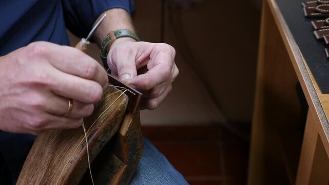 Close up shot of craftsman hands sewing a custom brown leather watch band with waxed thread and awl tool on a traditional wooden stitching pony in workshop.