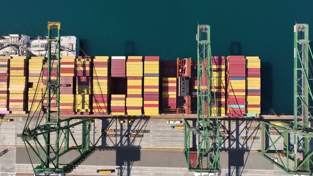 Cargo containers are stacked at a shipping terminal. Cranes are positioned for loading and unloading operations. Shipping congestion affects freight transportation. Delays impact global trade.