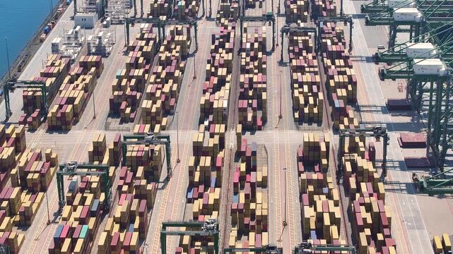 Containers are stacked at a busy port. Cranes move containers to and from ships. Delays are visible with congested cargo areas. Freight operations indicate high shipping volumes.