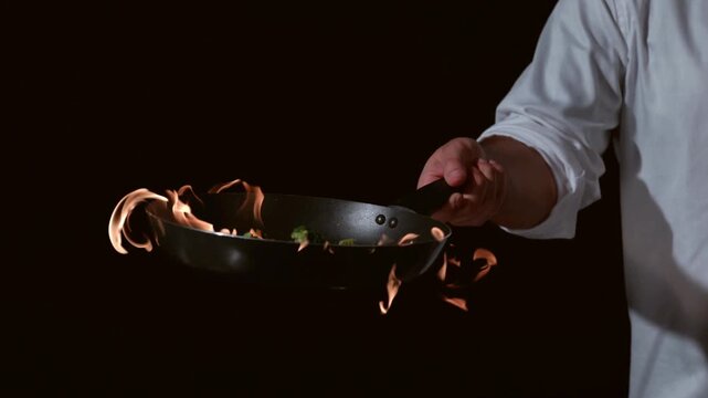 Professional chef tossing vegetables and shrimp in a flaming pan on a black background