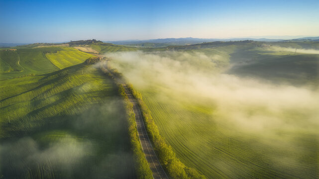 Aerial view of rolling green hills and a winding road shrouded in morning mist under a clear blue sky in Tuscany, Italy.