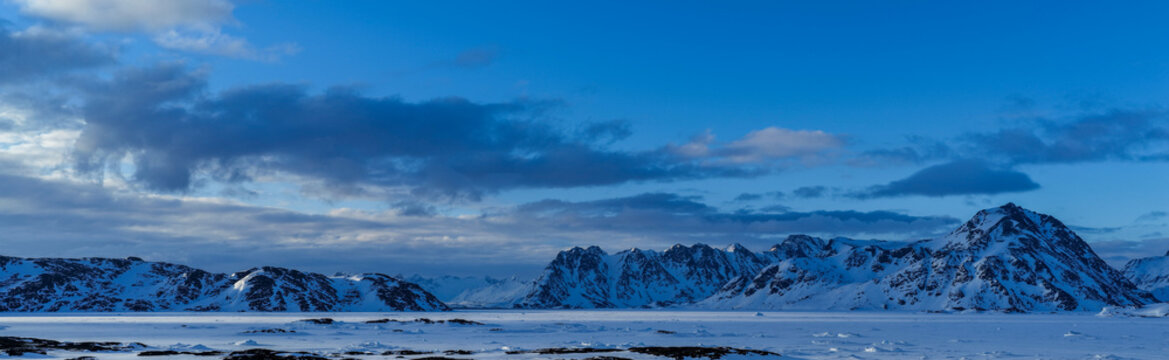 Aerial view of snow-capped mountain ranges and a vast frozen fjord under a deep blue sky with soft clouds Tasiilaq, Sermersooq, Greenland.