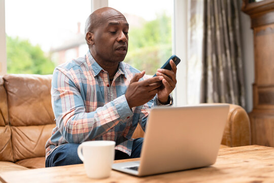 Man using phone while sitting on couch next to laptop with coffee cup on table during daytime at home