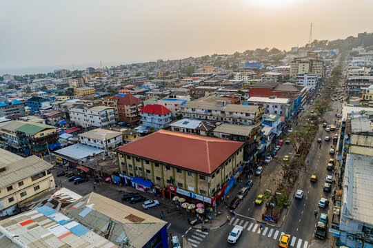 Aerial view of the dense urban landscape with colorful rooftops, busy streets with yellow taxis, and the coastline under a hazy sky in Monrovia, Montserrado, Liberia.