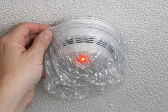 A hand holds a smoke detector wrapped in plastic, ready for testing or disposal