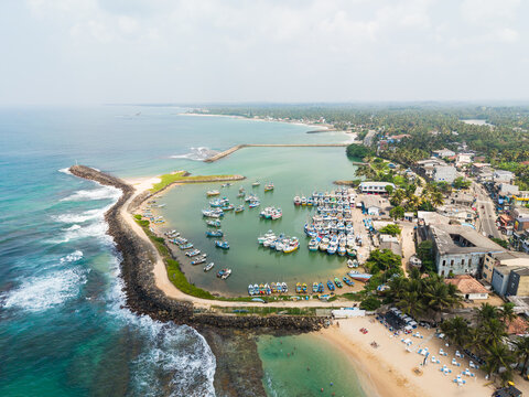 Aerial view of colorful fishing boats moored in the harbor protected by a stone breakwater along the sandy coastline in Hikkaduwa, Southern Province, Sri Lanka.