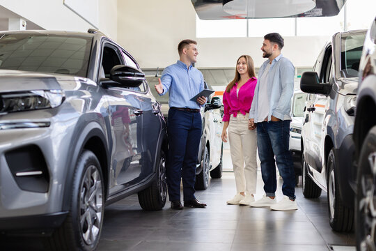 Young couple choosing a new vehicle at modern car dealership showroom while professional salesman presents automobile features and purchase options