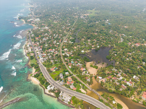 Aerial view of the coastline featuring Devil's Rock, lush tropical vegetation, and residential buildings along the road in Ahangama, Southern Province, Sri Lanka.