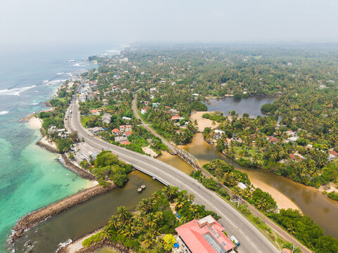 Aerial view of Devil's Rock with a coastal road bridge crossing a river surrounded by lush palm trees and turquoise ocean water in Ahangama, Southern Province, Sri Lanka.