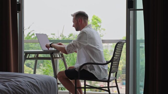 Man Sitting At Balcony Desk And Starting Remote Work Session In Hotel Workspace With Laptop And Smartphone, Flexible Work Lifestyle And Digital Nomad Routine